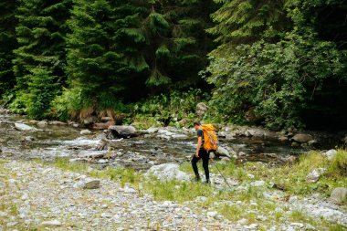 Sırt çantalı genç bir turistin nehir kenarındaki ormanın arka görüntüsü. Genç adam, turist teçhizatı ile donatılmış yürüyüş, yaşam tarzı. etkin yaşam modu, etkin dinlenme