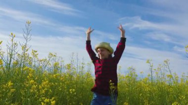 Küçük kız tarlaya atlayıp kolza tohumu tarlasında ellerini havaya kaldırdı. Mutlu enerjik çocuk uzun sarı çimenlerde zıpla