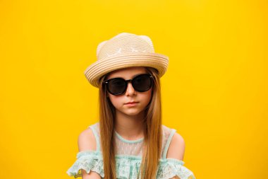Şık, ciddi, sarışın, güneş gözlüklü ve şapkalı bir kız. Kameraya bakan çocuk, sarı arka planda izole edilmiş bir video portresi. Stüdyo. Boşluğu kopyala