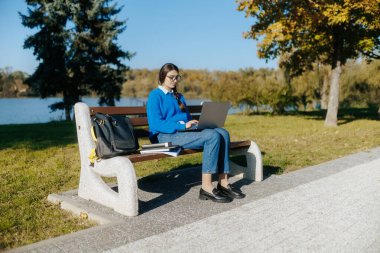 Sonbahar parkında dizüstü bilgisayarında gözlüklü ve mavi kazaklı genç bir kız. Kitapların yanında oturuyor, dijital öğrenme ile doğal sakinliği dengeliyor..