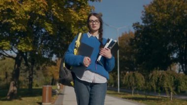 Odaklanmış genç bir öğrenci bir sonbahar parkında kitap, defter ve dizüstü bilgisayarını tutuyor. Sıcak bir şekilde giyinerek, dışarıda huzur bulur. Çalışmalarıyla doğayı dengeler..