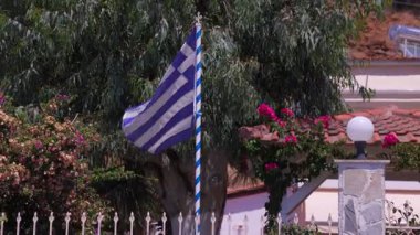 Mavi ve beyaz bayrak bir evin önünde dalgalanıyor. Bayrak direğin üzerinde ve bir bahçenin ortasında.