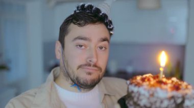 Bu görüntü, parti şapkası takan ve elinde yanan bir doğum günü pastası tutan orta yaşlı bir adamın evde tek başına kutlamasını gösteriyor..