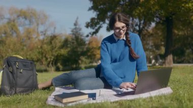 Gözlüklü genç bir kız öğrenci parkta çimlere oturur ve dizüstü bilgisayarıyla yazı yazar. O, kitaplarla çevrili ve sonbahar için sıcak giyinmiş olarak derslerine odaklanmıştır..