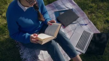 Bir sonbahar parkında açık havada kitap okuyan genç bir öğrencinin yakın çekimi. Dizüstü bilgisayarının yanındaki çimlerde oturur ve doğada huzurlu bir çalışma seansının tadını çıkarır..