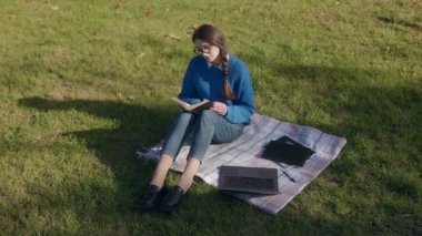 Sakin bir sonbahar ortamında, genç bir kız çimlerin üzerinde oturup kitap okuyor. Dizüstü bilgisayarı yanında yatıyor, açık havada çalışmanın özünü yakalıyor..