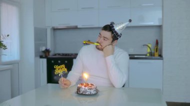 Parti şapkası ve günlük kıyafetleri olan orta yaşlı bir adamın sessiz bir mutfakta pastayla oturup mum yaktığı yalnız bir doğum günü kutlaması..