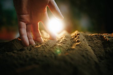 Bir el yere hafifçe dokunuyor, zengin toprakla etkileşime giriyor. Sıcak güneş ışığı arka planda yumuşak bir parıltı yaratıyor. Bu da doğayla yeryüzü arasındaki bağlantıyı gösteriyor..