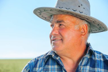 Güneşli bir yaz günü, kırsal kesimde tarım çiftliğinde dikilen şapkalı, neşeli, yaşlı bir erkek işçiyi kapatıyoruz.