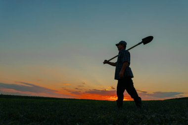 Çiftçilerin silueti, tarım endüstrisi kavramında kürekle yürüyor. Erkek gökyüzünün arka planında çarpıcı parlak renklerle. Günbatımında tarımda yürüyen insanlar. içinde