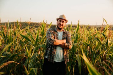 Hasır şapkalı bir çiftçi uzun mısır bitkilerinin arasında dururken gülümsüyor. Günbatımının sıcacık parıltısı kırsal hayatın güzelliğini vurgulayarak tarlaya huzur veren bir ışık saçıyor..