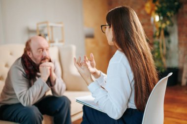 Beyaz tişörtlü bir terapist, bir müşteri terapi seansı sırasında düşüncelerini anlatırken dikkatle dinler. Oda sıcak ve davetkar, rahatlatıcı atmosferi arttırıyor..