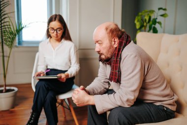 Gözlüklü ve beyaz bluzlu bir terapist terapi seansları sırasında derin düşüncelere dalmış gri kazaklı bir adamı dikkatle dinler..