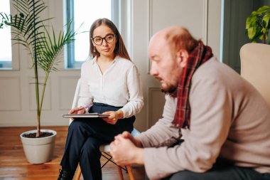 Bir terapist iyi aydınlatılmış bir ofiste müvekkilini dikkatle dinler. Terapist bir tablete not alırken müşteri dalgın görünüyor. Ortam sakin ve destekleyici..