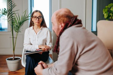 Bir terapist, iyi aydınlatılmış bir ofiste terapi seansı sırasında müşterisini dikkatle dinler. Çevre rahat, tartışma için rahatlatıcı bir atmosfer sağlıyor..