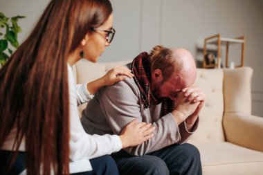 Genç bir kadın, rahat bir oturma odasında üzgün olan bir adamı teselli ediyor. Kadın, elini omzuna koyup ona ihtiyacı olduğunda empati ve özen göstererek onu rahatlatıyor..