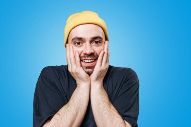 Bir adam coşkuyla tepki verir. Elleri yanaklarında gülümseyerek. Sarı bir bere takıyor ve koyu renk bir gömlek giyiyor. Neşeli mavi tavrını sergiliyor..