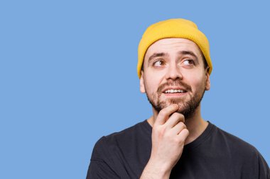Sarı bir bere takan genç bir adam, çenesini elinin üzerinde dinlendirirken düşünceli bir şekilde gülümsüyor, onun günlük davranışını vurgulayan sert bir mavi arka plan üzerine kurulmuş..