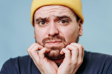 Sarı bereli genç bir adam sakalına dokunurken endişeli görünüyor. Yumuşak mavi arka plan, onun ifadesiyle çelişiyor. Endişe ve belirsizlik duygularını vurguluyor..