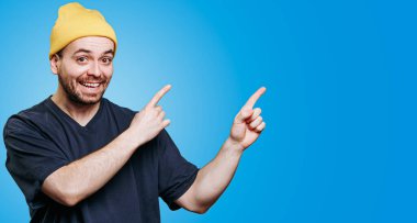 Parlak sarı bir bere takan neşeli bir adam büyük bir gülümsemeyle kenarları işaret ediyor. Arka planda canlı mavi bir renk var. Neşeli tavırlarını arttırıyor..