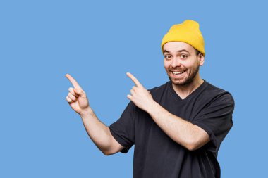 Sarı bereli ve koyu renk gömlekli bir adam enerjik bir şekilde her iki tarafı gösterirken gülümsüyor. Arka plan canlı bir mavi, neşeli ve canlı bir atmosfer yaratıyor..