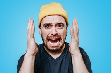 Parlak sarı bir bere takan bir adam, elleri çileden çıkmış bir şekilde hayal kırıklığına uğramış görünür. İfadesi, sert mavi bir arka plana karşı güçlü bir duygu taşıyor..