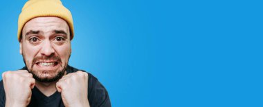 Sarı bereli bir adam heyecanını yumruklarını sıkarak ve gülümseyerek gösterir. Parlak mavi arka plan coşkulu ifadesini ve enerjisini vurguluyor..