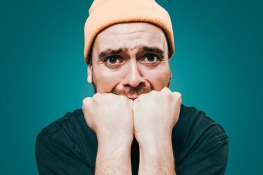 Bir adam yumruklarını sıkarak ve endişeli bir bakış atarak kaygılarını ifade eder. Sarı bir bere takıyor ve sade bir arka plana karşı ciddi bir tavrı var..