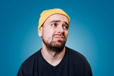 Sarı bereli ve koyu renk gömlekli bir adam yüz ifadesinden hayal kırıklığına uğradığını ifade eder. Arkaplan pürüzsüz, canlı bir mavi, anı canlandırıyor..