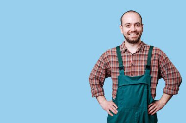 Yeşil tulumlu ve ekose gömlekli neşeli bir adam elleri kalçasında, basit mavi bir arka plana karşı sıcak bir gülümseme sergiliyor..