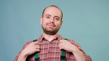Ekose gömlek giyen ve yeşil tulumlu neşeli bir adam kendinden emin duruyor. Arkadaşça bir ifadesi var.