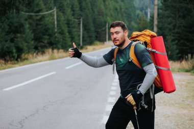 Solo gezgin, sakin bir yolda yürür, heyecanı göstermek için başparmağını kaldırır. Bereketli orman ve berrak gökyüzü, doğal güzelliklerin arasında aktif bir yaşam tarzını gösteriyor..