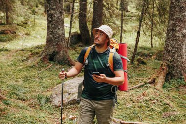 Sakallı bir yürüyüşçü, verimli bir ormanlık alanı keşfederken bir akıllı telefon tutar ve teknoloji ile doğanın karışımını gösterir. Cihazı huzurlu arazide gezinmek için kullanıyor..