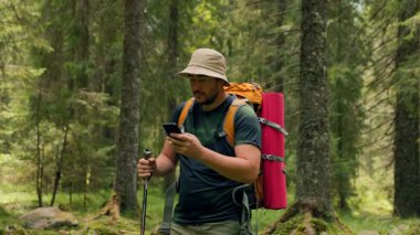 Sakin bir ormanda, genç bir Latin gezgin elinde akıllı bir telefonla duruyor, teknolojiyi doğayla harmanlıyor. Sırt çantası ve yürüyüş direği var, keşfetmeye hazır..