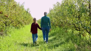 Bir anne ve kızı canlı bir elma bahçesinde el ele yürürler, bereketli yeşillik ve ağaçlarla çevrili iken sıcak güneşin altında taze elma toplarlar..