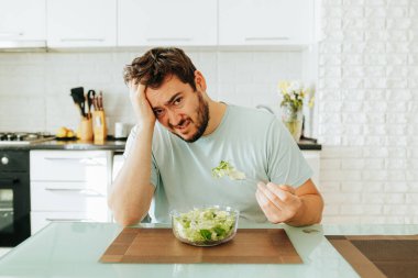 Genç bir adam, bir kase salata yerken gözle görülür şekilde hayal kırıklığına uğramış, çağdaş bir mutfakta bir masada oturuyor. Mutfak iyi aydınlatılmış, temiz ve düzenli bir ortam sunuyor..