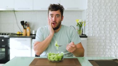 Şaşırmış bir ifadeyle mutfak masasında oturan bir adam bir çatal dolusu salata yer. Parlak mutfakta beyaz dolaplar ve önünde bir kase taze yeşillik var..