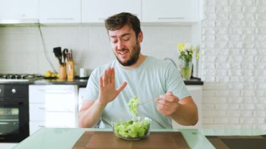 Bir adam modern bir mutfak masasında otururken salatadan hoşlanmadığını ifade eder. Oda aydınlık, sağlık odaklı çağdaş bir tasarım sergiliyor..