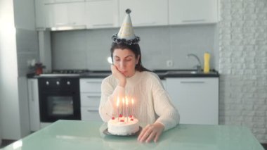 Orta yaşlı bir kadın, sıradan giyinmiş, doğum gününü pastanın üstünde mumlarla kutluyor, temiz bir mutfakta oturuyor, düşünceli bir bakışla hayatı yansıtıyor..