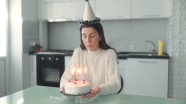 Düşünceli genç bir kadın doğum gününü evde tek başına kutluyor. Modern mutfağında otururken yanan mumlarla pastaya bakıyor..