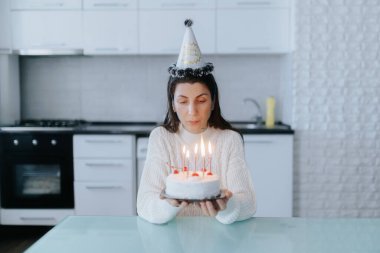 Rahat bir kazak giymiş düşünceli bir kadın doğum gününü yalnız kutluyor. Modern mutfağında mum ışığında bir pasta, sessiz bir yalnızlık ve yansıma sahnesi..