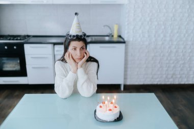Beyaz giyinmiş üzgün, düşünceli bir kadın doğum gününü mum ışığında bir pastayla kutluyor. Modern bir mutfak masasında sessizce oturuyor ve yalnızlığı simgeliyor..