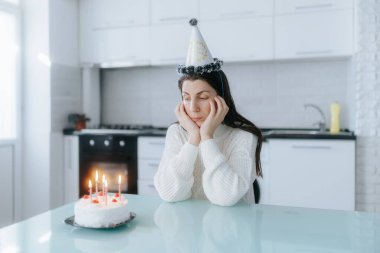 Yalnız bir kadın mutfak masasında elinde doğum günü pastası ve mumlarla oturuyor, parti şapkası takıyor, sessiz bir kutlama sırasında duygularını yansıtıyor..