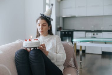 Beyaz bir kadının, oturma odasının sakin atmosferiyle çevrili bir kanepede yalnız başına pasta ve mumlarla doğum gününü kutladığı duygusal bir sahne..