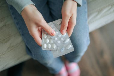 Kanepede oturan biri elinde iki paket ilaç tutuyor. Elleri plastiği kavrıyor ve rahat bir ortamda dalgın dalgın duruyorlar..