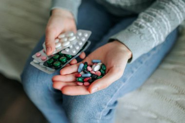 Bir insan kanepeye oturur ve elindeki renkli hapları sınıflandırır. İlaç paketleri görüntülenir, farklı şekil ve boyutlarda bir dizi ilaç sergilenir.