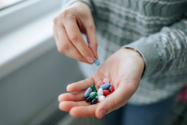 Bir insan sabah rutini sırasında bir avuç dolusu renkli tabletten hapları seçiyor. Güneş ışığı pencereden süzülür, sahneyi aydınlatır.