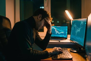 Bir adam gecenin geç saatlerinde karanlık bir ofiste birden fazla bilgisayar ekranında finansal verileri incelerken, işinin zorluklarını yansıtan bir stres yaşar..