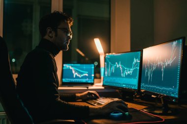 Odaklanmış bir birey gece geç saatlere kadar çalışır. Masa lambasıyla aydınlatılan şık ve modern ofis ortamında birden fazla ekranda finansal piyasa verilerini analiz eder..
