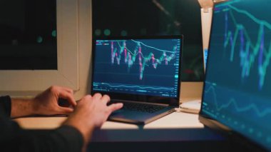 Eller bir dizüstü bilgisayar ve ikinci bir monitörde borsa çizelgelerinde gezinir. Görünüm, karanlık sonrasında piyasa eğilimlerini ve dalgalanmaları gösteren ayrıntılı grafikleri yakalar.